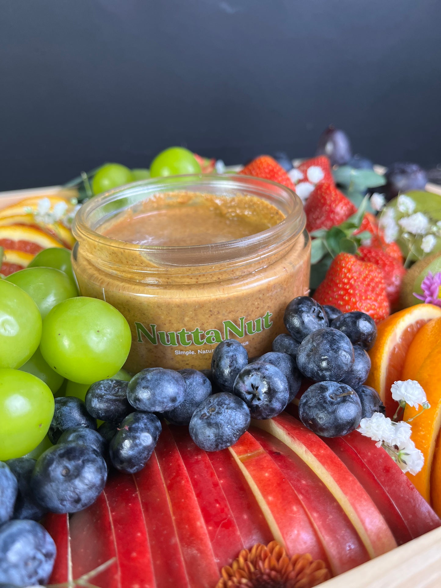 Jar of Nutta Nut spread surrounded by fruits and flowers on a dark background