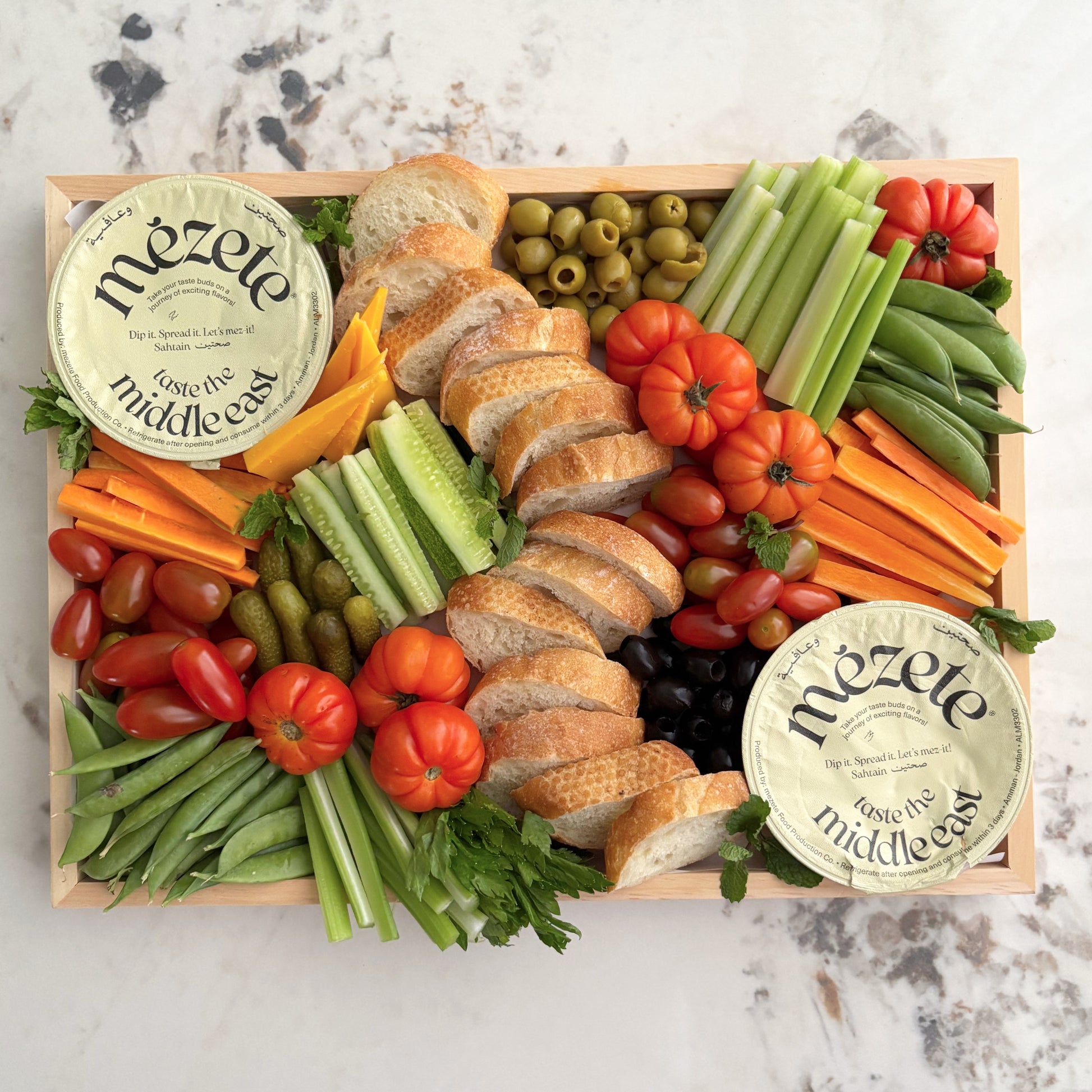 Mezete Hummus crudite board with bread, vegetables, and olives on a marble surface. Platter of assorted vegetables, bread, and two types of dip on a marble surface. Le Bliss Platter’s Double Hummus Crudités Platter available in Jakarta & Surabaya.