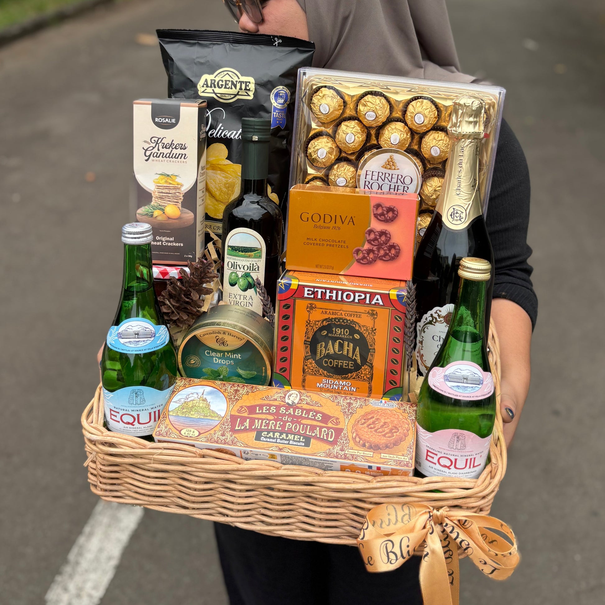 Person carrying a large gift basket filled with snacks and drinks on a street. Gift basket with snacks, drinks, and chocolates on a dark surface. hamper jakarta, hamper surabaya, gourmet hamper jakarta