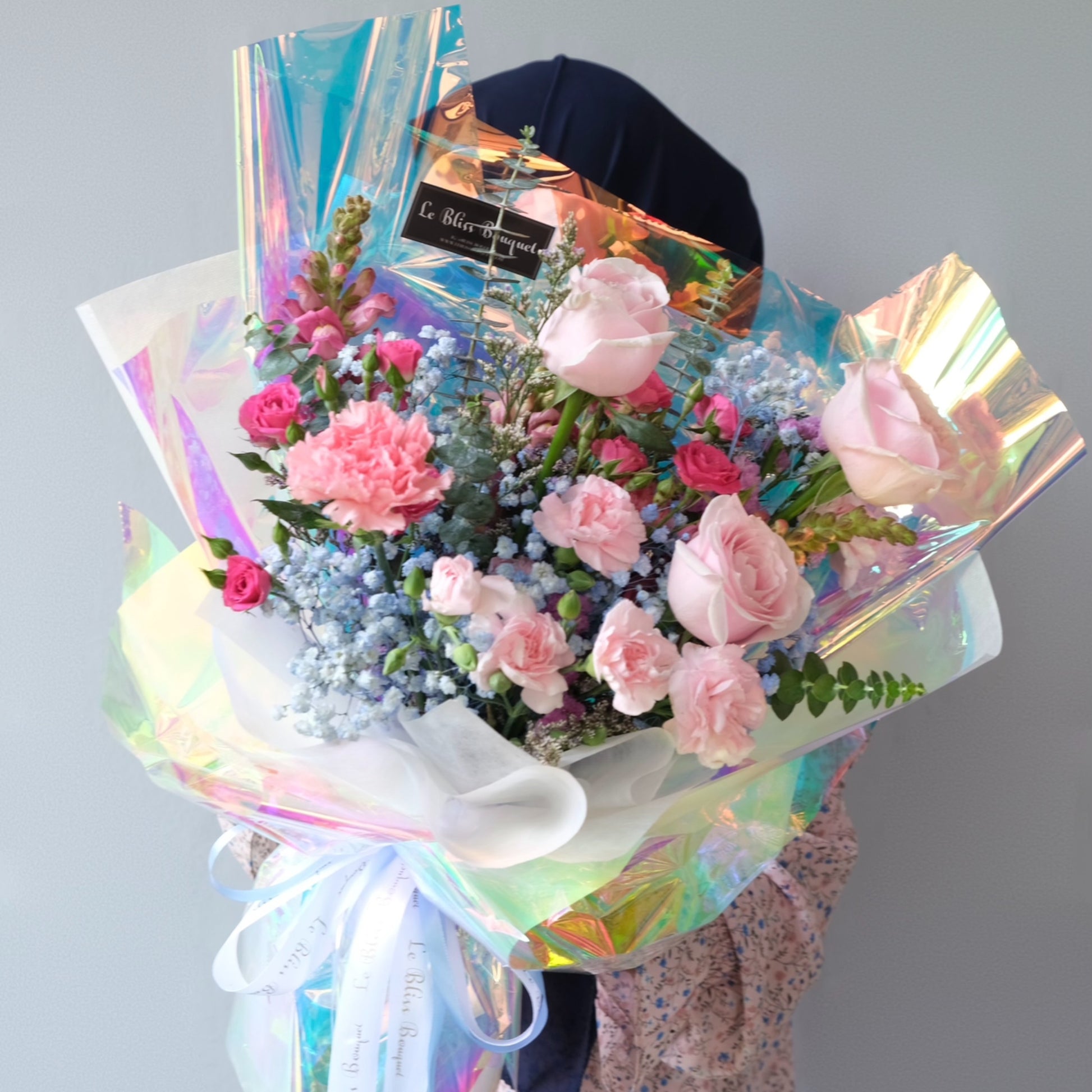 Person holding a bouquet of flowers wrapped in iridescent cellophane against a plain background.  Le Bliss Bouquet’s Pink Hologram Bouquet — trendy pink roses wrapped in holographic paper. Same-day delivery to Jabodetabek, Surabaya & Cirebon.