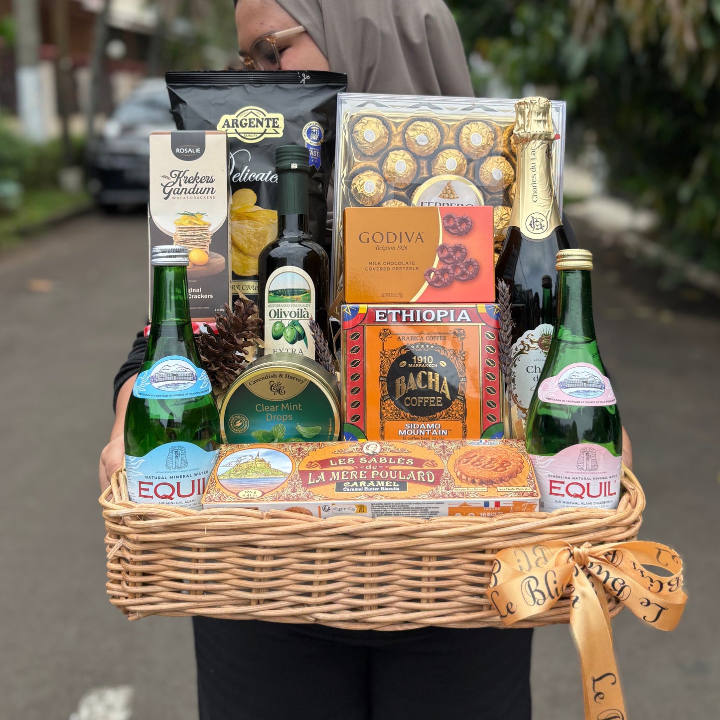 Person holding a gift basket with various items including bottles and boxes on a street.. Wicker basket with snacks, drinks, and chocolates on a stool in an urban street setting. hamper jakarta, hamper surabaya, gourmet hamper jakarta