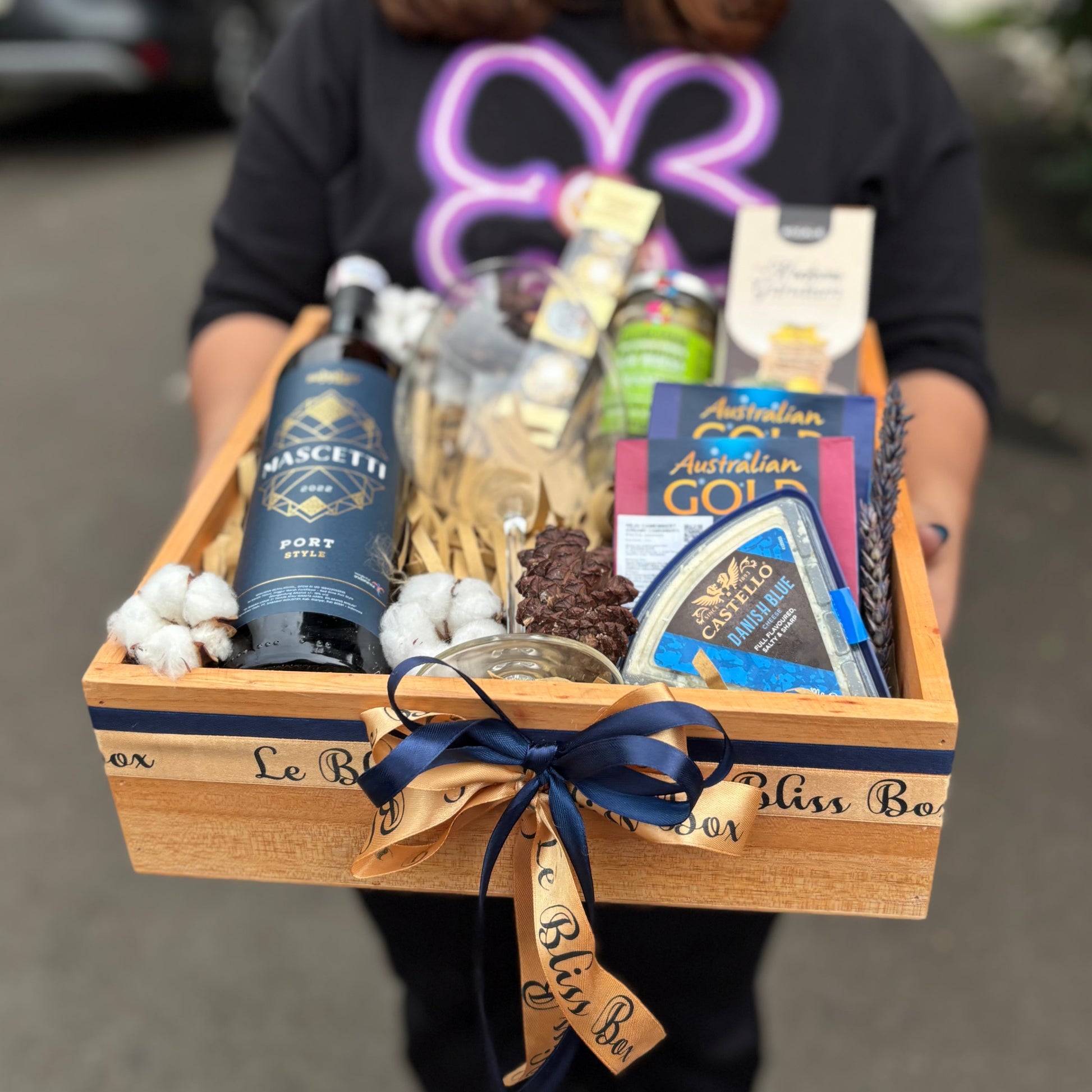Person holding a gift basket with various items on a street background. Wine Hamper Jakarta, Surabaya, Indonesia. Gift basket with wine, cheese, and other snacks on a dark surface. By Le Bliss Box