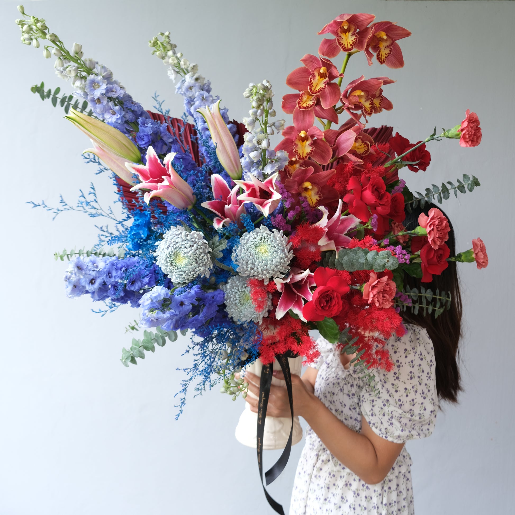 Person holding a large, colorful bouquet of flowers against a plain background. vase delphinium blue with lily and red cymbidium by le bliss bouquet. florist jakarta, florist surabaya, florist cirebon, toko bunga jakarta