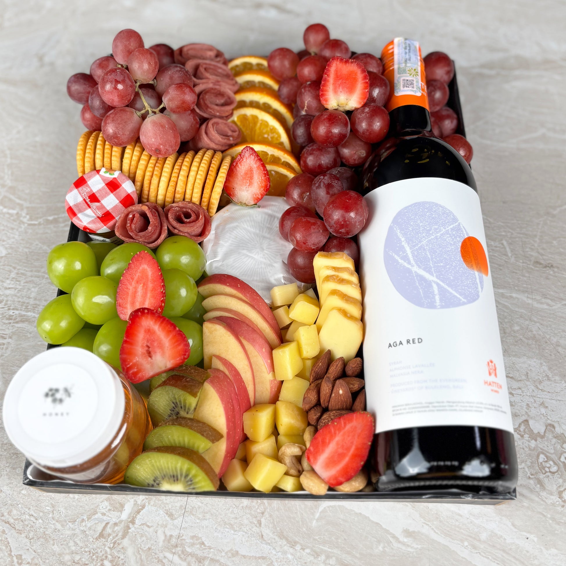 Platter of fruits, cheeses, and meats with a bottle of wine on a light gray surface. Charcuterie board with fruits, cheeses, and a bottle of wine on a light gray surface. Red Wine Cheese Platter with artisanal cheeses, fresh fruits, honey, jam & smoked beef. 