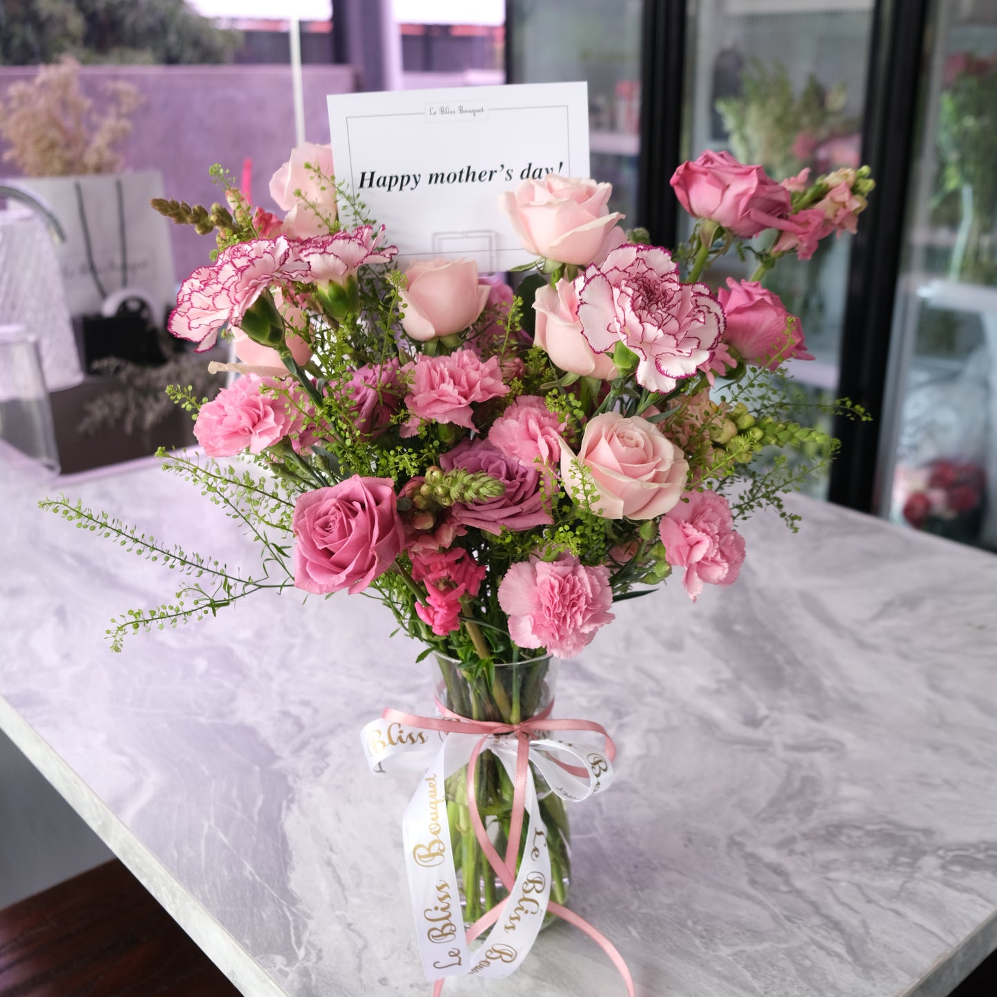 Vase of pink flowers with a card on a marble surface. Pink Sonata Flower Vase dari Le Bliss Bouquet. Kado Hari Ibu yang elegan dengan bunga premium. Pengiriman Surabaya, Jakarta & Cirebon. Perfect Mother’s Day flower gift. 