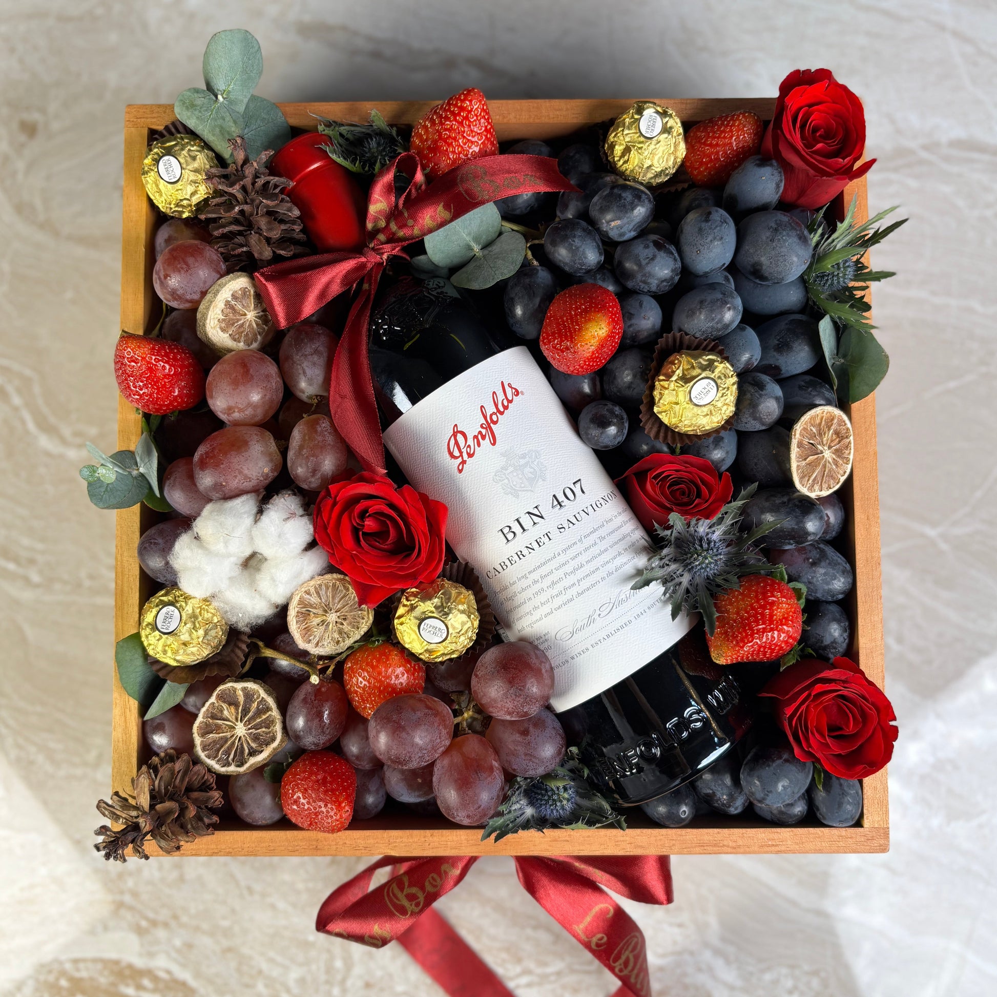 Wine bottle in a decorative box with fruits, flowers, and candies on a light background. Gift basket with fruits, flowers, and a bottle of wine on a textured surface. Penfolds Wine Hamper Jakarta, Surabaya. With Fresh autumn grapes, red globe grapes and strawberries and chocolate. Wine Hamper Jakarta. Christmas hamper jakarta