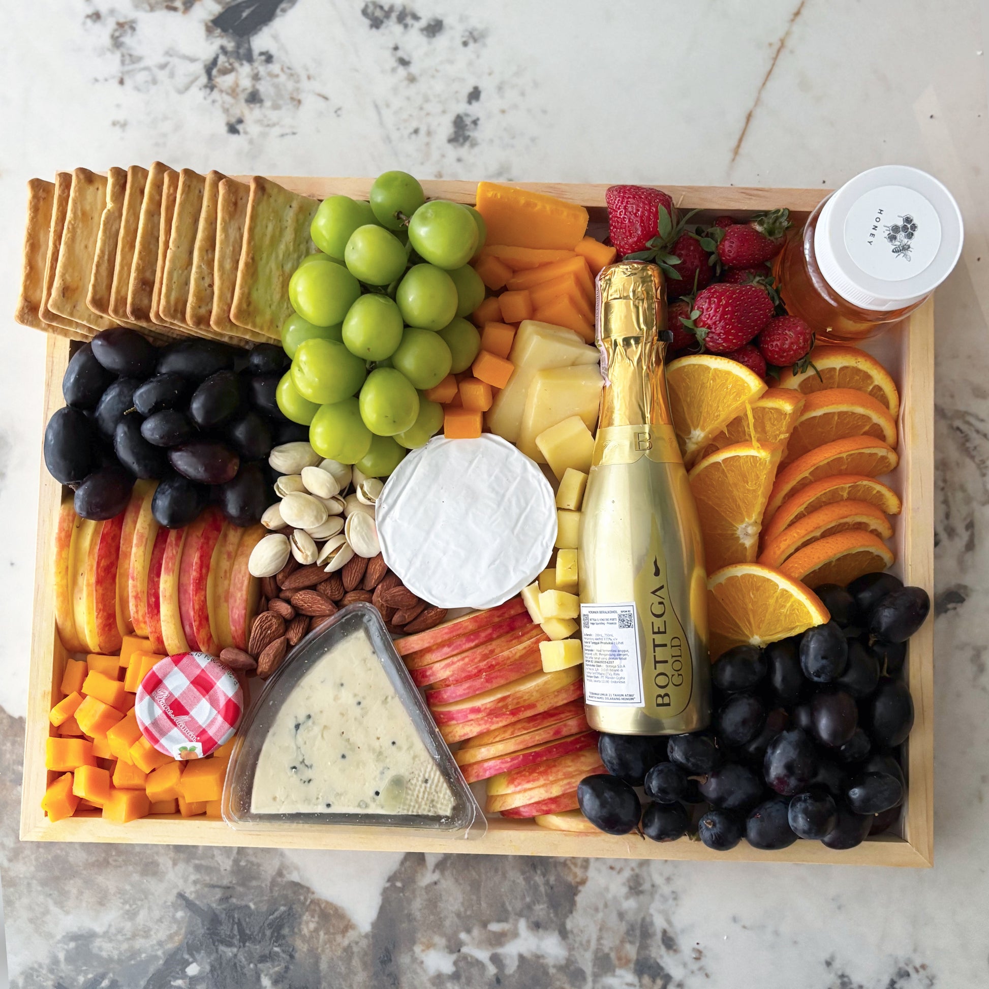 Wooden tray with assorted fruits, cheeses, and a bottle of champagne on a marble surface. best cheese platter jakarta, best cheese platter surabaya