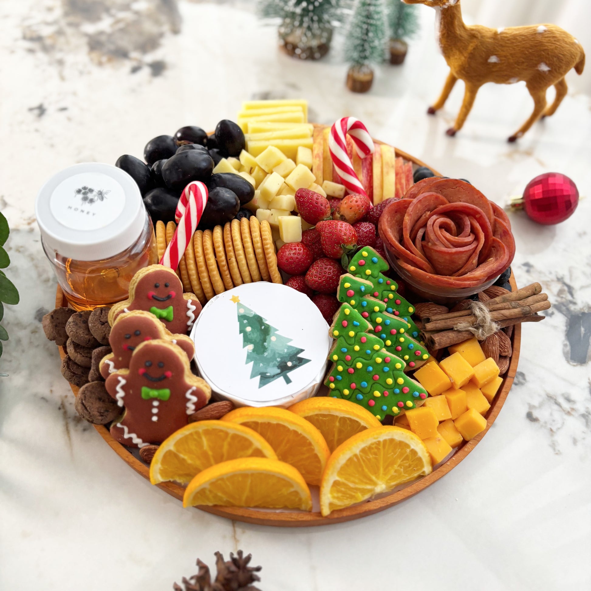 christmas charcuterie board surabaya with fruits, cookies, and a small Christmas tree on a table with festive decorations.  festive charcuterie board with fruits, cheese, and meats on a marble surface. Christmas Round Cheese Platter | Christmas Platter Jakarta & Surabaya – Le Bliss Platter