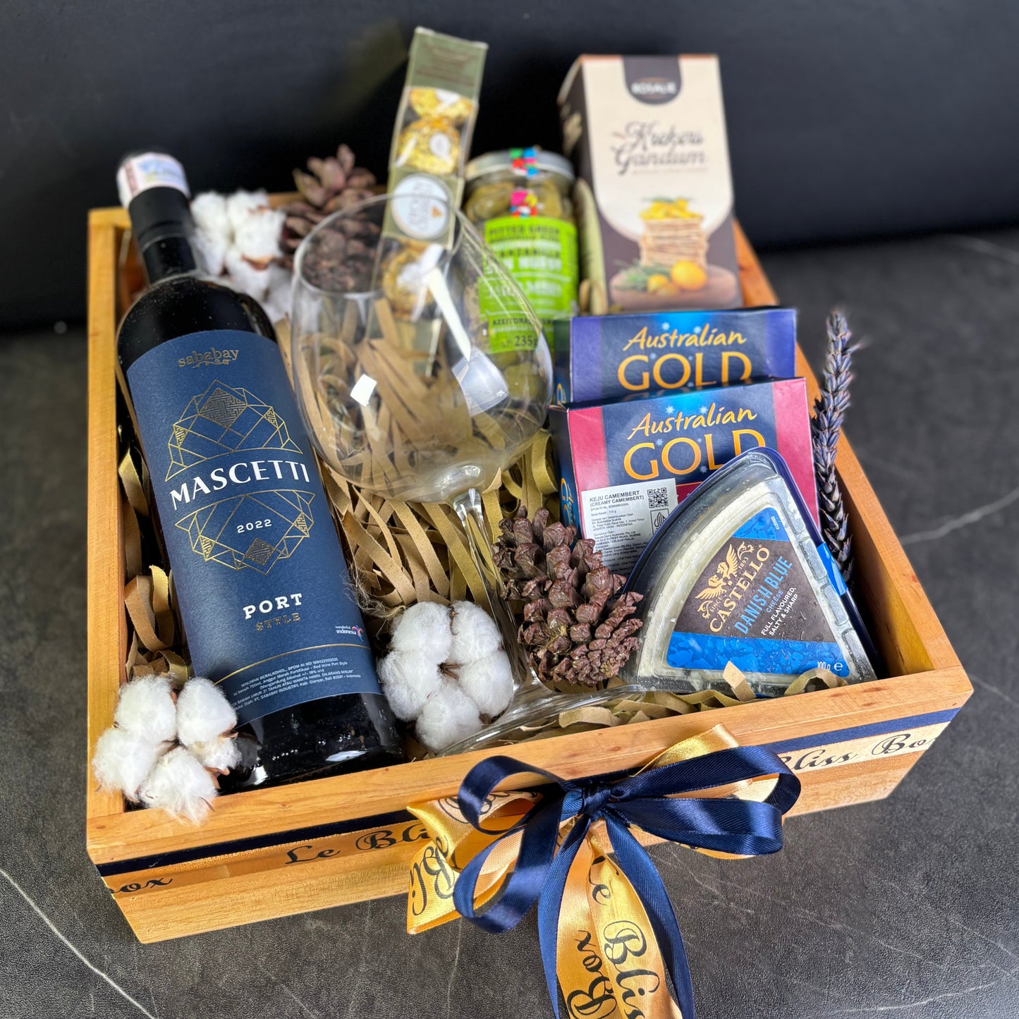 wooden basket with wine, cheese, and other snacks on a dark surface. Wine Hamper Jakarta, Surabaya. Gift basket with wine, cheese, and other snacks on a dark surface. By Le Bliss Box